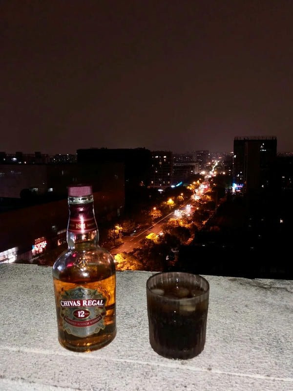 Người đàn ông lớn tuổi ngồi một mình bên bàn nhậu đơn sơ, chỉ có chai bia và vài món ăn, trong một không gian tĩnh lặng.