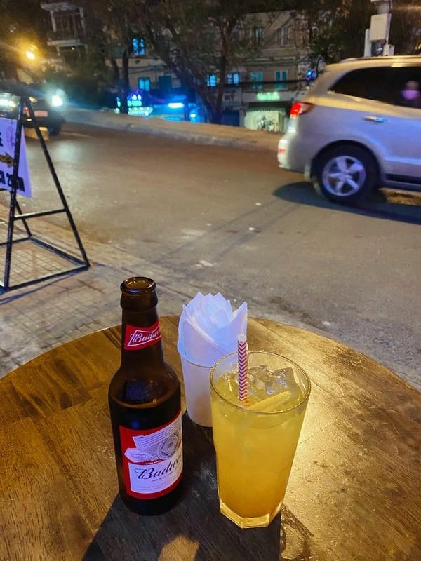 Bàn tay cầm ly bia đặt trên bàn, ánh đèn mờ ảo tạo không gian riêng tư, thể hiện sự cô độc.